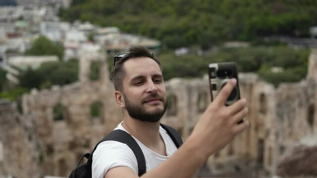 Young adult tourist with phone takes selfie with Acropolis hill, ruins of ancient Athens. Traveler in journey trip photographs old Greece, shoots pictures of himself, temples and shrines tourism.