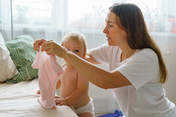 Mommy dressing little girl in dress. Naked baby in diaper standing by sofa and looking at camera