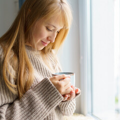 Happy woman with cup of hot drink standing next to window in sweater. Slow-paced lifestyle