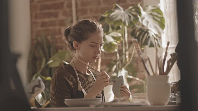 Chest up slowmo shot of young Caucasian woman in apron painting handmade ceramic bowl during group masterclass in cozy pottery workshop