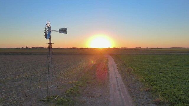 Aerial pan shot capturing rustic water pumping windmill spinning with the wind in the middle of fertilised grass field during sunrise in the early morning.