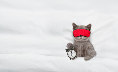 Cozy tabby kitten wearing mask for sleep shows alarm clock on a bed at home. Top down view. Empty...