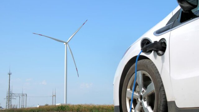 Windmill generates energy under blue sky. Electric vehicle with connected charger stands against rotating machine near power lines