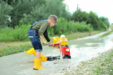 Little boy plays with toy cars in a puddle on a dirt road. - obrazy, fototapety, plakaty