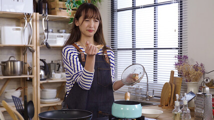 asian housewife trying a small amount of soup on stove at home kitchen, making bad face for its...