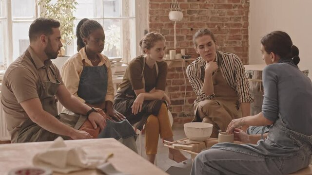 Slowmo stab shot of small multiethnic group of young people sitting around pottery wheel in workshop looking at female potter giving masterclass