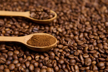 Close-up of wooden spoons with ground coffee and roasted coffee beans on the background of scattered fresh roasted coffee grains with copy space for advertisement.