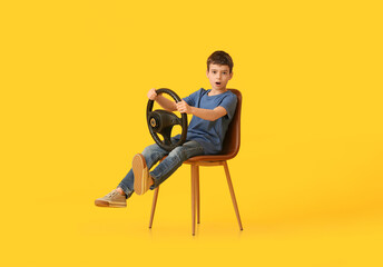 Shocked little boy with steering wheel sitting in chair on color background