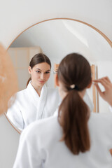 Young woman applying natural essential oil with pipette on her face near mirror