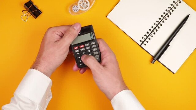 A successful man financier or economist in a white shirt is counting on a small calculator on a bright yellow background with a notepad, a proofreader and a paper clip