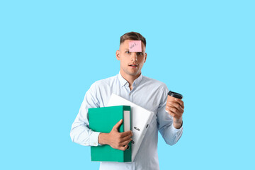 Young man with folders, sticky note with word WORK on his forehead and coffee against color background