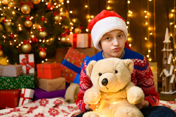 Fototapeta na wymiar Child girl posing in new year or christmas decoration. Festive lights and lots of gifts, an elegant Christmas tree with toys. The girl is wearing a red sweater and a Santa hat, she playing with bear