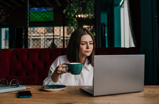 a focused brunette businesswoman in a white shirt is sitting at a table with a cup of coffee and a laptop