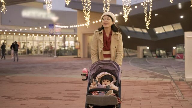 cheerful asian mother taking evening walk with her baby daughter in the pushchair outside a shopping center with Christmas light during the winter holiday