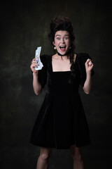 Creative portrait of young excited woman in image of medieval royal person in black dress isolated on dark vintage background.