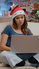 Bored adult using laptop and looking at movie on television for christmas eve celebration. Woman wearing santa hat with device, watching TV and feeling lonely on holiday festivity.