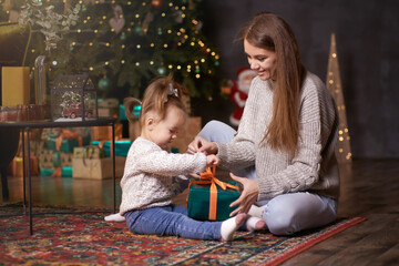 Happy mother with daughter near Christmas tree unpacks present. Having fun together. Young family...