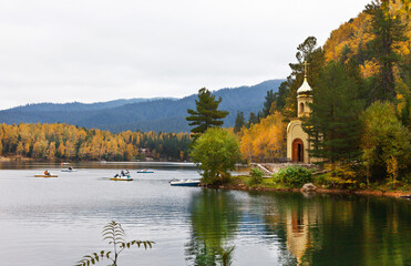 Obraz premium Tourists love to relax in the fall on the shore of a quiet warm Emerald Lake, surrounded by a beautiful multi-colored yellowed forest, ride water bikes and visit a small Orthodox church on the shore