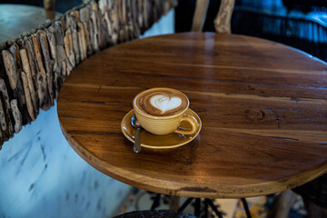 Trendy yellow cup of hot cappuccino on wooden table background. Heart shape latte art for symbol of love.