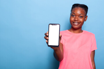 Portrait of african american teenager holding smartphone with white empty screen in hand, showing...