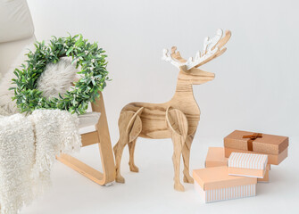 Wooden reindeer, gift boxes, armchair with plaid and Christmas wreath on light background, closeup