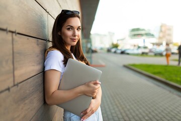 Photo of pretty attractive beautiful smiling positive charming young brunette young woman holding gray computer laptop wearing stylish clothes in white t-shirt and light blue jeans in the street in