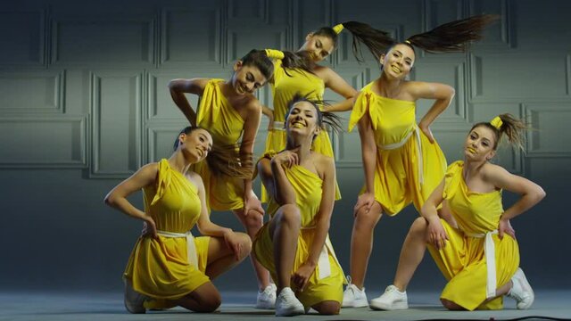 Group of beautiful , funny, stylish girls in yellow dresses dancing inside pavilion with grey background  . Synchronized dance of female ensemble for video clip  Woman dancers with the same outfit 