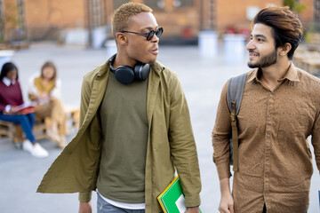 Multiracial male students walking and talking on territory of university campus. Concept of education and learning. Idea of student lifestyle. Young serious indian and black guys with notebooks