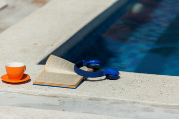 orange cup, book, blue earphones near swimming pool