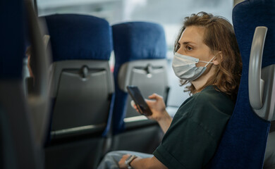Young woman in a mask on her face sitting in a train with a smartphone in her hands, traveling using the railroad, safe travel during a pandemic