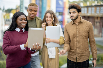 Multiracial students watching something on digital tablet outdoors. Concept of education. Remote and e-learning. Idea of student lifestyle. Smiling young friends at university campus