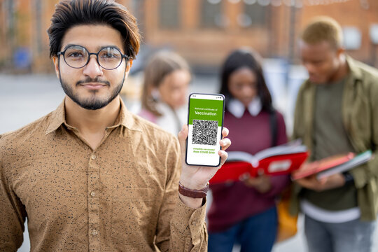 Indian student guy show smartphone with app of Coronavirus Vaccination with background of friends at university campus. Concept of education and learning. Health protection during Coronavirus pandemic