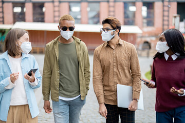 Multiracial students walking on territory of university campus. Concept of education and learning. Idea of students lifestyle. Young guys and girl in medical mask. Health protection during pandemic