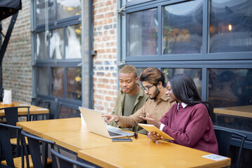 Fototapeta premium Multiracial students watching something on laptop while sitting at table outdoors. Concept of remote and e-learning. Students lifestyle. Focused young indian man, black girl and guy. University campus