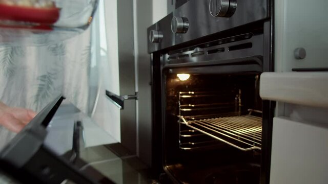 Chef wearing beige tracksuit puts transparent baking sheet inside preheated and illuminated modern oven in kitchen closeup slow motion