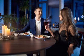 Charming woman and handsome man looking at each other while sitting together at table and drinking red wine. Romantic atmosphere at home for happy young couple.