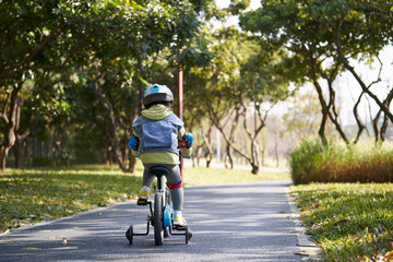 rear view of asian little girl riding bike outdoos in park - obrazy, fototapety, plakaty