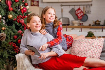 Obraz na płótnie Canvas Two joyful sisters are sitting at home near the christmas tree and looking at tablet monitor