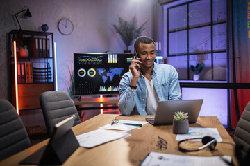 Young african man using wireless laptop and talking on smartphone while sitting at desk. Company member using modern gadgets for work at office.