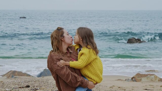 Young loving mom is holding her small daughter in her arms, sharing love and care, they kiss each other and look at camera, outdoor leisure on beach, Foreground, Slow motion. 