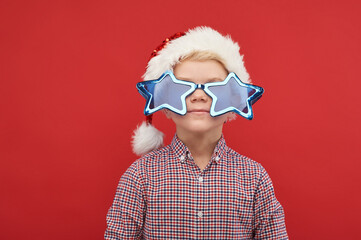 Happy child wearing Santa Claus hat and carnaval sunglasses. Portrait of funny kid against red background