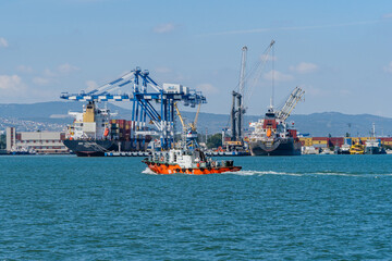 Fototapeta premium Yellow tug PILOT is heading towards embankment. Container ships Horizon, MSC Tasmania Panama are at berths under loading. Novorossiysk commercial sea port. Novorossiysk, Russia - September 15, 2021