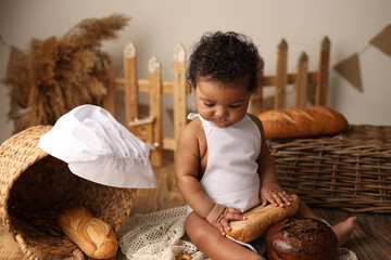 a cute dark-skinned kid with curly hair in a chef's costume has a bagel and a roll. High-quality photography