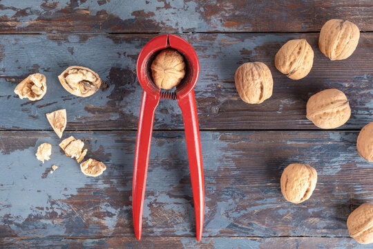 A bright red heavy metal nutcracker with a walnut inside, next to a scattering of inshell whole walnuts and a few chopped nuts, against a textured wooden background with cracked paint.