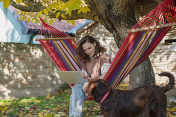 Obraz na płótnie Canvas Young happy woman freelancer with laptop and dog in hammock at backyard of country house on autumn day