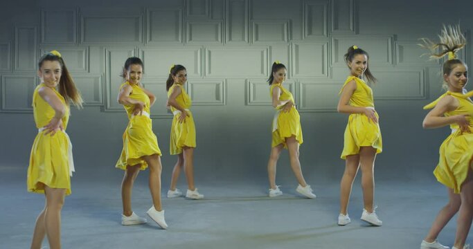 4k Group of beautiful , funny, stylish girls in yellow dresses dancing inside pavilion with grey background  . Synchronized dance of female ensemble for video clip  Woman dancers with the same outfit 