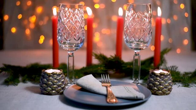 Festive table served with grey natural linen tablecloth, glasses for champagne, plate, fork, napkin on background of four burning red festive Christmas candles and shining bokeh garland lights