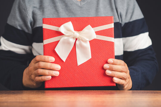Man in sweater holding a red gift box on a wooden table. A parcel wrapped in red paper and tied with a white ribbon. Close-up photo. Concept of Christmas, New Year, Anniversary, Happy birthday, etc