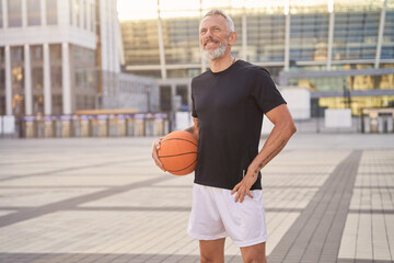 Active middle aged man in sportswear looking away, standing outdoors with basketball ball, ready...