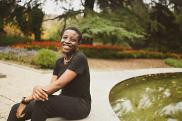 Attractive happy smiling young natural beauty short haired African woman wearing total black laughing sitting on the edge of a fountain in nature summer park